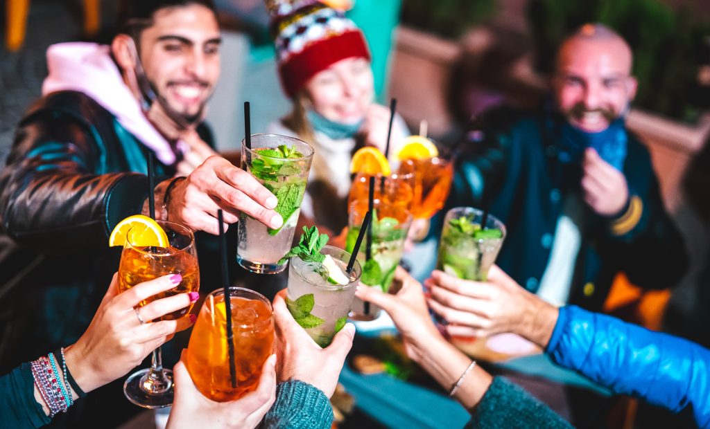 Happy friends toasting drinks at night bar with open face mask - New normal nightlife concept with milenial people having fun together on winter clothes at apreski - Vivid filter with focus on glasses